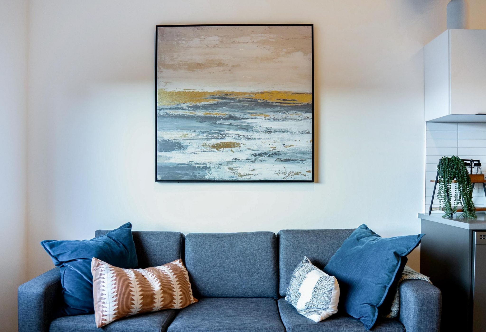 A modern gray sofa with blue and patterned pillows sits below an abstract seascape painting on the wall.