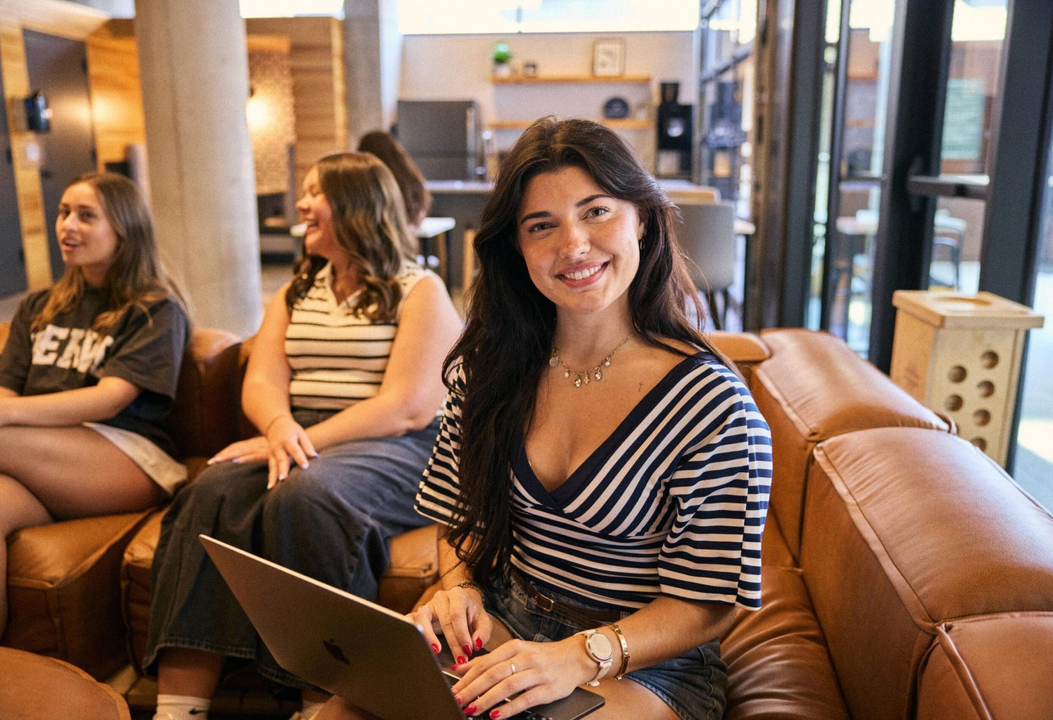 A young woman with dark hair using a laptop while sitting on a couch with others in a shared workspace.